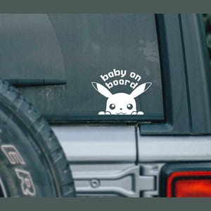 Könnte beinhalten: Weißer Autoaufkleber mit einem Pikachu-Cartoon-Charakter und dem Text "baby on board". Der Aufkleber befindet sich auf der Heckscheibe eines Fahrzeugs.