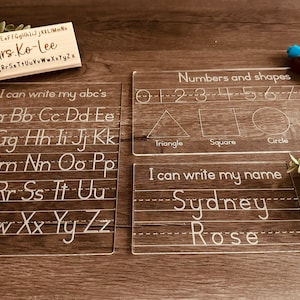 May include: Clear acrylic learning boards with printed letters, numbers, and shapes. One board says "I can write my abc's" and another says "I can write my name" with the name "Sydney Rose". A wooden nameplate is also visible.