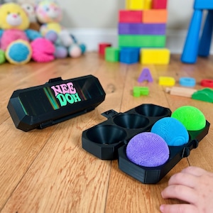Op de afbeelding: Een zwarte plastic opbergkoffer met de tekst Nee Doh op het deksel, vergezeld van een bijpassend zwart dienblad met drie gestructureerde, bolvormige stressballen in de kleuren paars, felblauw en neongroen.