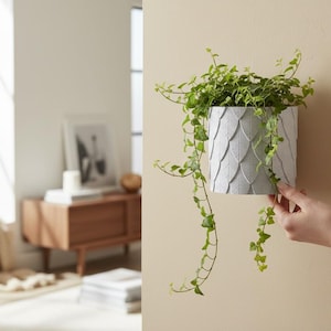 Puede incluir: Una maceta de pared gris texturizada con un patrón de hojas, llena de hiedra verde colgante. La maceta está montada en una pared beige. La imagen está bien iluminada, mostrando el verde vibrante de la planta contra el fondo neutral.
