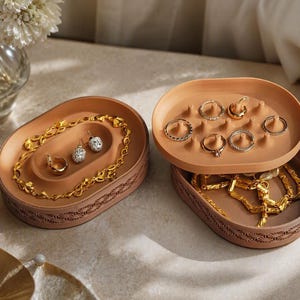 May include: Two oval-shaped, terracotta-colored jewelry organizers with intricate geometric patterns along the sides. One organizer features a tiered design with a top tray containing small pegs for rings, while the other holds a gold chain and earrings.