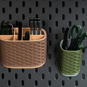 May include: A tan, oval-shaped desk organizer with six compartments containing pens and tools, alongside a smaller, cylindrical olive green pen holder containing scissors, both featuring a repeating geometric star pattern on their exterior.