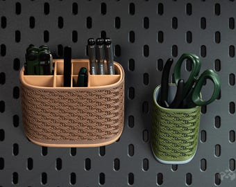 May include: A tan, oval-shaped desk organiser with six compartments containing pens and tools, alongside a smaller, cylindrical olive green pen holder containing scissors, both featuring a repeating geometric star pattern on their exterior.