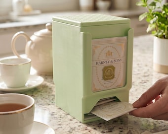 May include: A light green tea bag dispenser with a textured lid, holding a stack of Harney & Sons tea bags. The vertical rectangular container features an open front slot for easy access to individual tea bags, with one bag partially pulled out.
