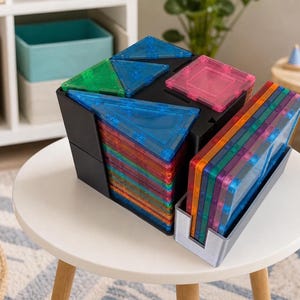 May include: A black plastic storage bin filled with translucent, colorful magnetic building tiles in square and triangular shapes, accompanied by a separate gray plastic tray holding a stack of additional square magnetic tiles.