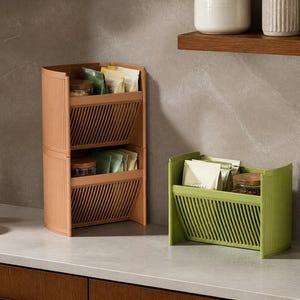 May include: A stackable storage organizer in terracotta orange and a single unit in sage green. Each unit features a slatted front panel, open top, and curved side walls, designed to hold small items like tea bags, jars, and packets on a counter.