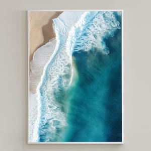 May include: An aerial view of a wave crashing on a sandy beach. The water is a vibrant turquoise blue, and the wave is white with foam. The sand is a light beige color.