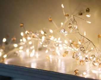 Puede incluir: Una cadena de luces blancas cálidas con cuentas de vidrio transparentes y ámbar. Las luces están enhebradas en un alambre plateado y son perfectas para agregar un toque de brillo a cualquier habitación.