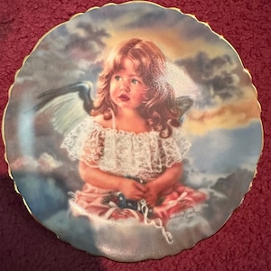 May include: Decorative plate featuring a young girl with angel wings, seated on a cloud. The girl wears a white lace top and a pink skirt. The plate has a scalloped edge with a gold trim and a signature in the bottom right corner.