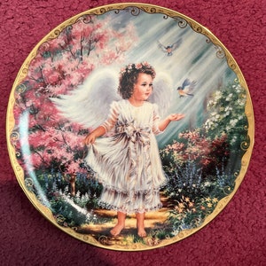 以下が含まれることがあります： 白いドレスと大きな翼を着けた若い天使が、ピンクの花と2羽の青い鳥がいる庭にいる装飾皿。お皿には金の縁取りがあります。
