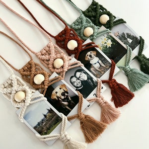May include: A collection of macrame photo holders in various colors, including cream, pink, brown, and green. Each holder features a wooden bead and a tassel, designed to display square photographs. The holders are arranged on a white surface.