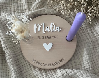 May include: A wooden plaque with the name Malia, the date December 28, 2023, and the text "Wie schön, dass du geboren bist!"  A white heart and a purple candle are also on the plaque.