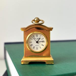 May include: A small gold-toned metal mantel clock with a rounded top and a circular handle. The clock face is white with black Roman numerals, black hands, and the text "W. Widdop Est. 1883" and "QUARTZ" printed in the center.