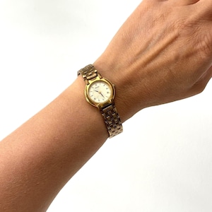 May include: A gold-tone analogue wristwatch featuring a round white dial with thin gold hands and hour markers. The watch has a metallic link bracelet with a textured pattern and a secure clasp, providing a classic look for daily timekeeping.