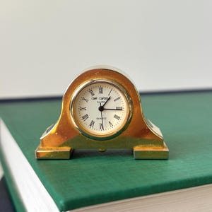 May include: A gold-toned mantel clock with an arched frame and a white circular face. The clock features black Roman numerals, black hands, and the text "Sir Williams Est. 1883" and "QUARTZ" printed in black on the center of the dial.