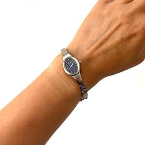May include: A silver-toned wristwatch featuring an oval-shaped case and a dark blue dial with the brand name Pulsar printed in white. The watch has a slender, polished metal link bracelet that secures around the wrist with a clasp mechanism.