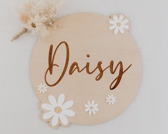 Peut inclure: Une pancarte ronde en bois avec le nom "Daisy" gravé en lettres brunes. La pancarte est décorée de fleurs de marguerite blanches.