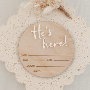 May include: A wooden round sign with the text "He's here!" in white lettering. The sign has blank lines for name, time, date, weight, and length.