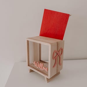 Pode incluir: Uma caixa de madeira clara com tampa vermelha e ranhura superior. Apresenta um laço rosa e o nome "Willow" em escrita rosa. A caixa tem um design moderno e é feita de madeira clara.
