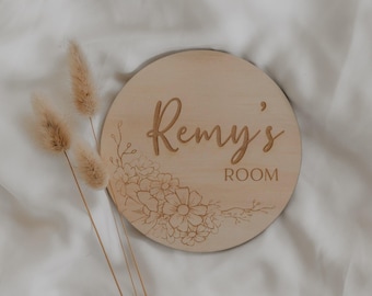 Peut inclure: Panneau rond en bois clair avec l'inscription "Remy's ROOM" en écriture cursive. Décor floral gravé dans le coin inférieur gauche. Le panneau est posé sur une surface blanche.