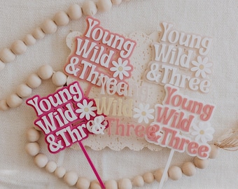 May include: Four pink, white, and yellow cake toppers with the text "Young Wild & Three" and a daisy design.