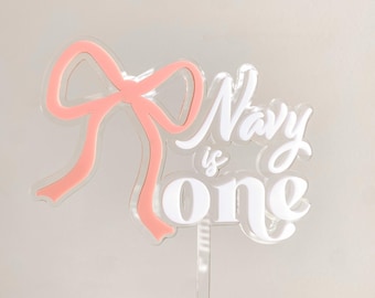 May include: White acrylic cake topper with the text "Navy is one" in a cursive font. A pink bow is attached to the left side of the topper.