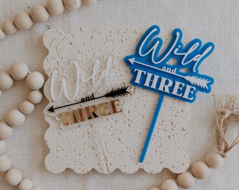 以下が含まれることがあります： 「Wild and THREE」の文字が入った2つのバースデーケーキトッパー。1つは白、もう1つは白い矢印が付いた青色です。3つ目のバースデーケーキトッパーは金色です。トッパーは、木製のビーズと紐が付いた、質感のあるオフホワイトの表面にあります。