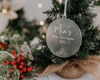 Könnte beinhalten: Ein silberner Weihnachtsschmuck mit dem Text "Mia's First Christmas 2023" und einem Weihnachtsbaum-Design. Der Schmuck hängt an einem kleinen Weihnachtsbaum mit Jute-Basis, umgeben von festlichem Grün und roten Beeren.