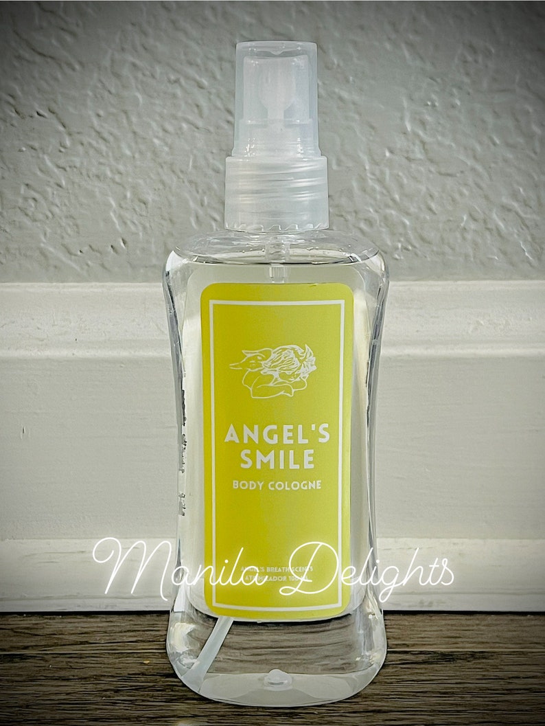 May include: A clear glass bottle of Angel's Smile body cologne. The bottle has a yellow label with a white angel design and the text "Angel's Smile Body Cologne".