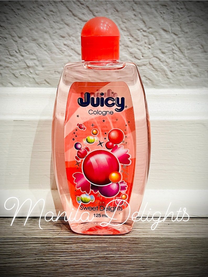 May include: A clear glass bottle of Juicy Cologne with a red cap and a pink and white label. The label says "Juicy Cologne" and "Sweet Delights" with a design of colorful candy and bubbles. The bottle contains 125 ml of cologne.