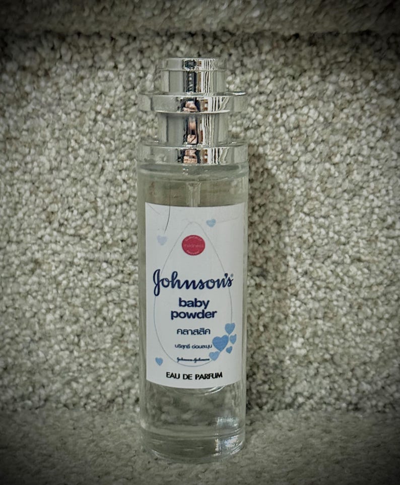 May include: A clear glass bottle of Johnson's baby powder fragrance with a silver cap and a white label with blue hearts. The label reads "Johnson's baby powder  粉末  วิทย์  douche  Eau de Parfum".