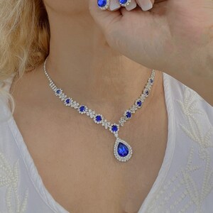 May include: A silver necklace with a teardrop pendant set with blue rhinestones. The necklace is paired with matching earrings with teardrop blue rhinestone accents.