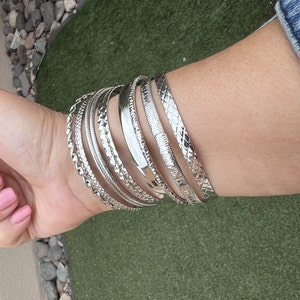 Op de afbeelding: Een verzameling zilveren armbanden met verschillende texturen, waaronder gladde, gevlochten en patroonontwerpen. De armbanden zijn op een pols gestapeld en tonen hun diverse stijlen en reflecterende oppervlakken.