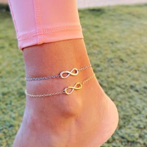 May include: Two infinity symbol anklets, one silver and one gold, on a person's ankle. The anklets are delicate and dainty.