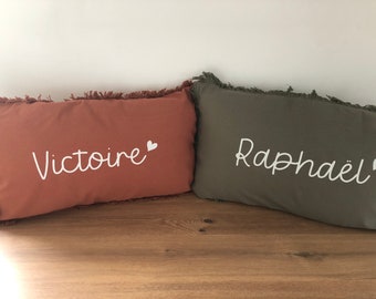 May include: Two decorative throw pillows with fringed edges. The pillow on the left is a burnt orange colour with the name "Victoire" embroidered in white with a small white heart. The pillow on the right is a muted olive green with the name "Raphaël" embroidered in white with a small white heart.