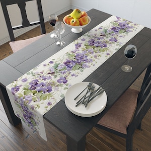 Puede incluir: Un camino de mesa blanco con un diseño floral con flores moradas y verdes. El camino de mesa está colocado sobre una mesa de madera oscura con dos copas de vino, un cuenco de manzanas y una pila de platos blancos con cubiertos.