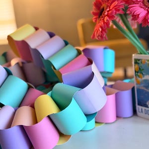 May include: A colorful paper chain in pastel shades of pink, yellow, blue, and purple. The chain is arranged in a wavy pattern, with a vase of pink gerbera daisies in the background.