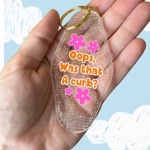 May include: A clear plastic keychain with glitter and pink flowers. The keychain has the text "Oops, Was that A curb?" in orange letters.