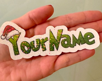 Może przedstawiać: Personalizowana naklejka z napisem Your Name wykonanym zieloną, teksturowaną czcionką z brokatowym wykończeniem. Litera Y jest zwieńczona czerwono-białą czapką Mikołaja. Naklejka ma białą obwódkę i mieniący się, holograficzny wygląd.
