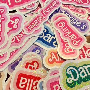 May include: A collection of colorful, glittery stickers with names written in white lettering. The names include Sabrina, Parking, Jess, Dan, Ella, and Skinny Water.