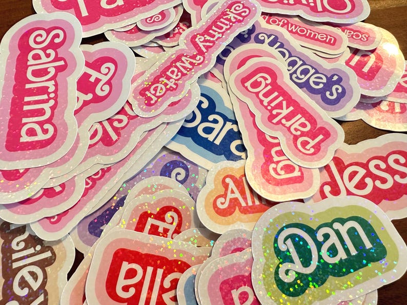 May include: A collection of colorful, glittery stickers with names written in white lettering. The names include Sabrina, Parking, Jess, Dan, Ella, and Skinny Water.