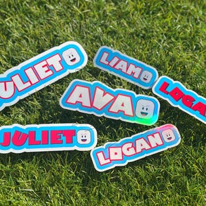 May include: Several name stickers with a cartoon face design are displayed on a green grassy surface. The names include "Juliet", "Liam", "Ava", and "Logan", each in a different color scheme with a blue outline.