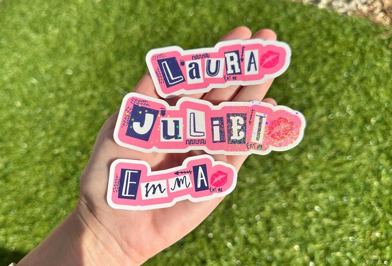May include: Three pink and white stickers with the names "Laura", "Juliet", and "Emma" written in black letters. Each sticker has a pink lip design and the words "Eat Me" written in black letters.