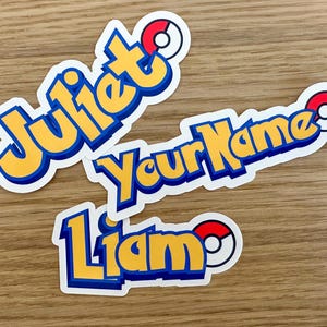 May include: Three colorful stickers with names written in a bold, cartoon font. Each sticker features a red and white Pokeball graphic. The names on the stickers are Juliet, Your Name, and Liam.