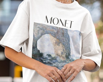 Può includere: Maglietta bianca con una stampa grafica di un dipinto di Claude Monet. Il dipinto raffigura una scogliera rocciosa con un arco e il mare. Il testo "MONET" è stampato sopra il dipinto.