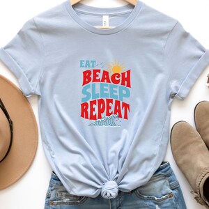 May include: Light blue t-shirt with the text "Eat, Beach, Sleep, Repeat" in red and blue lettering. The shirt features a sun and a wave graphic.