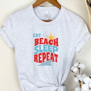May include: A grey t-shirt with the text "Eat Beach Sleep Repeat" in red, blue and yellow. The text is in a retro font style with a sun and waves graphic.
