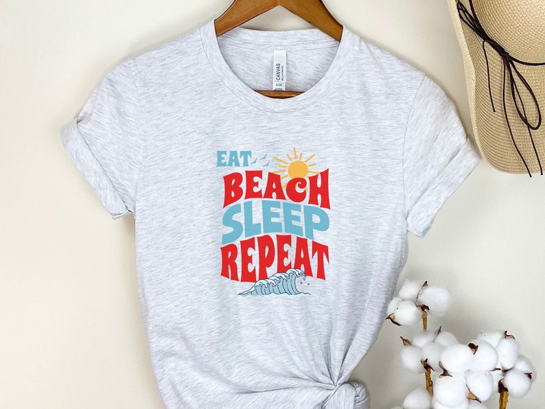 May include: A grey t-shirt with the text "Eat Beach Sleep Repeat" in red, blue and yellow. The text is in a retro font style with a sun and waves graphic.