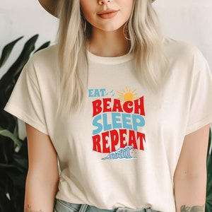 May include: A white t-shirt with a retro-style graphic that says "Eat Beach Sleep Repeat" in red, blue, and yellow. The graphic also includes a sun, birds, and a wave.