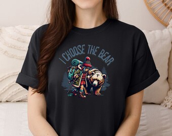 May include: Black t-shirt with a graphic of a girl wearing a hat and carrying a backpack while walking with a bear. The text "I choose the bear" is printed on the shirt.